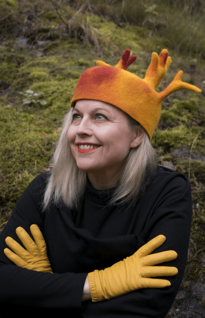 Elina Saari istuu hirviä-viikonloppu kurssia varten tehdyn oranssisävyisen hatun kanssa sammalleista kivikkoa vasten.