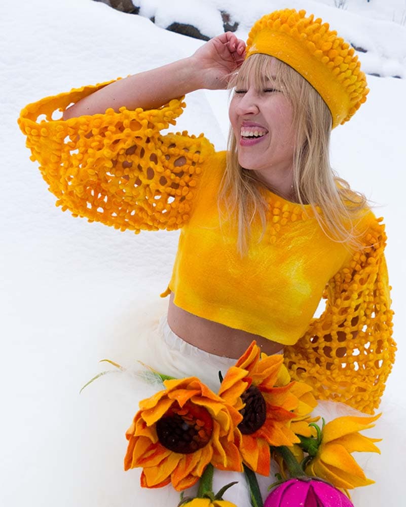 Kuvassa nainen huovutettu nappulapaita ja hattu päällä hangessa, sylissä huovutettuja auringonkukkia.